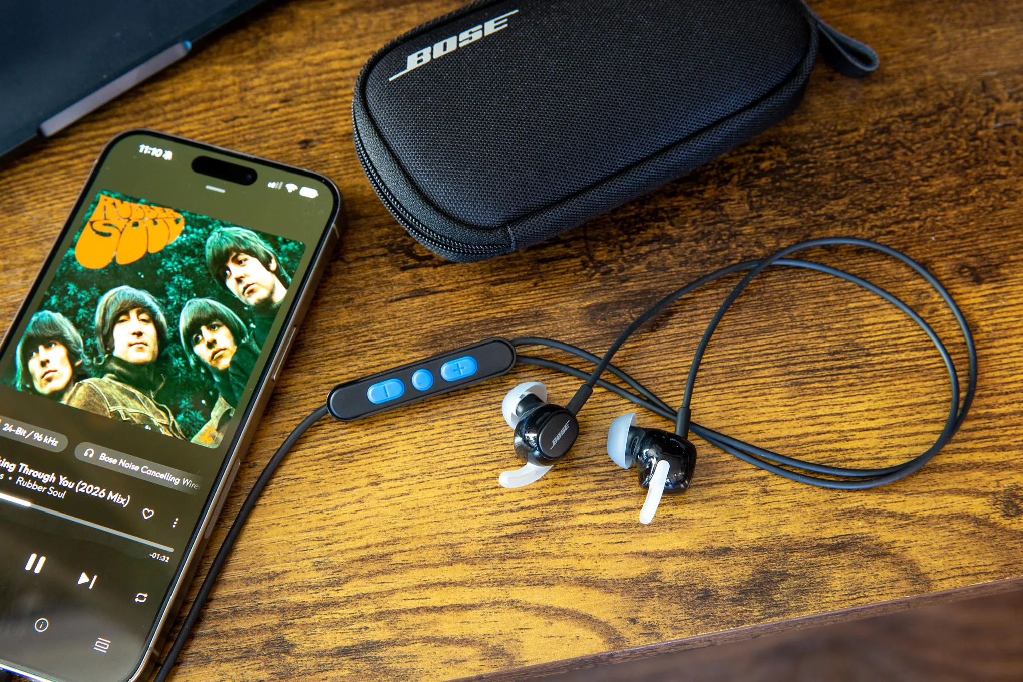 The Bose Noise Cancelling Wired Earbuds on a wooden desk next to their case, an iPhone, and a MacBook.