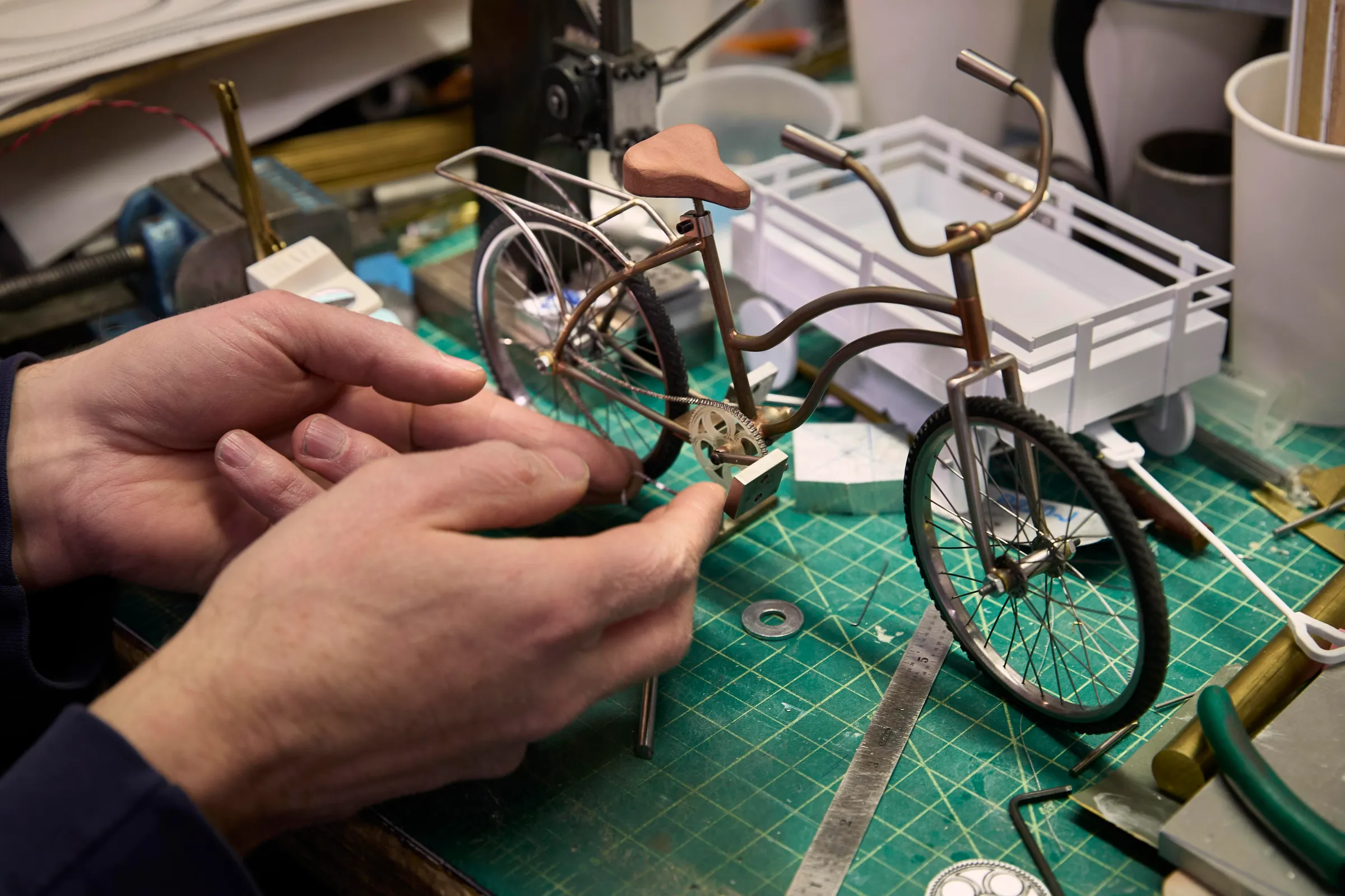 A pair of hands working on a model of a bicycle on a crafting table, with various tools and parts nearby.