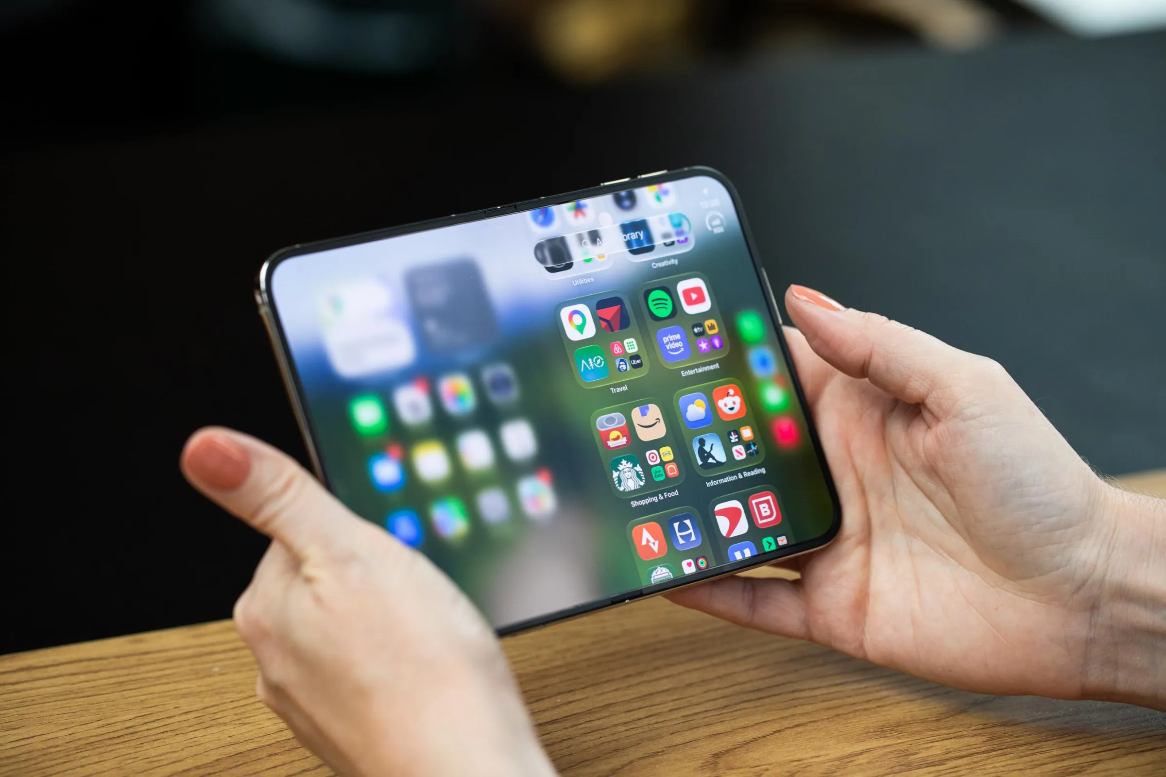 Hands holding the iPhone Duo open to the inner screen in landscape orientation. The left side of the screen is blurred out, while the right side shows the app library.