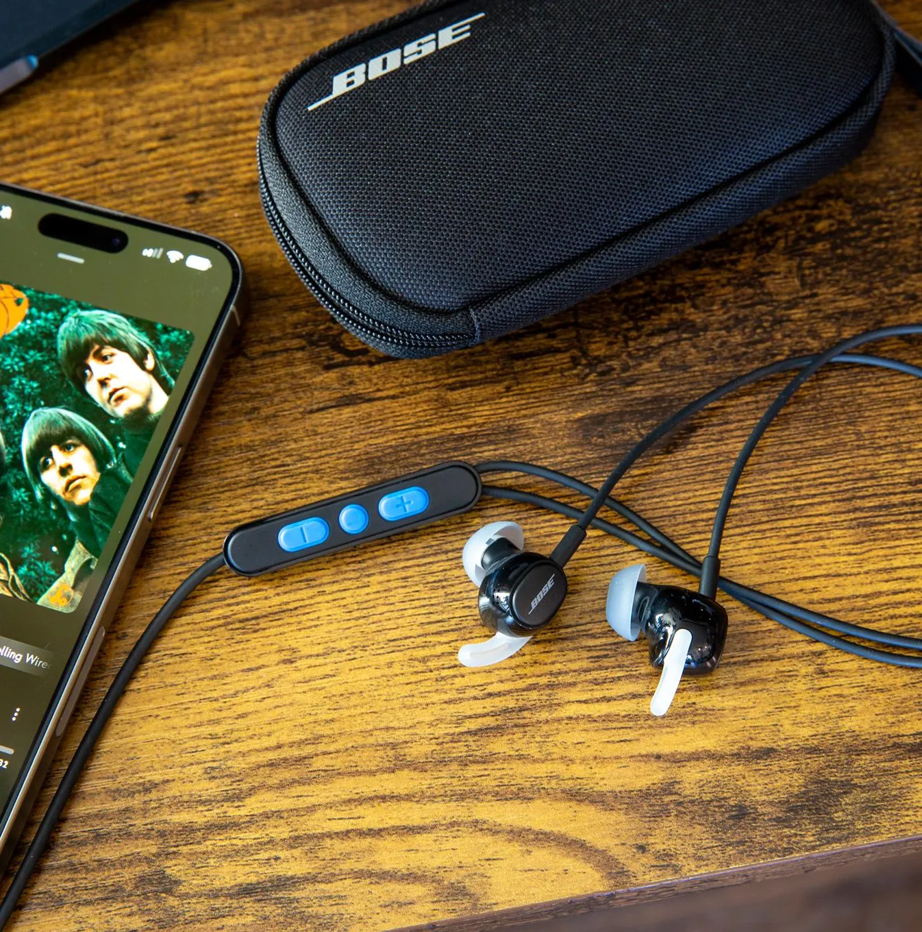 The Bose Noise Cancelling Wired Earbuds on a wooden desk next to their case, an iPhone, and a MacBook.