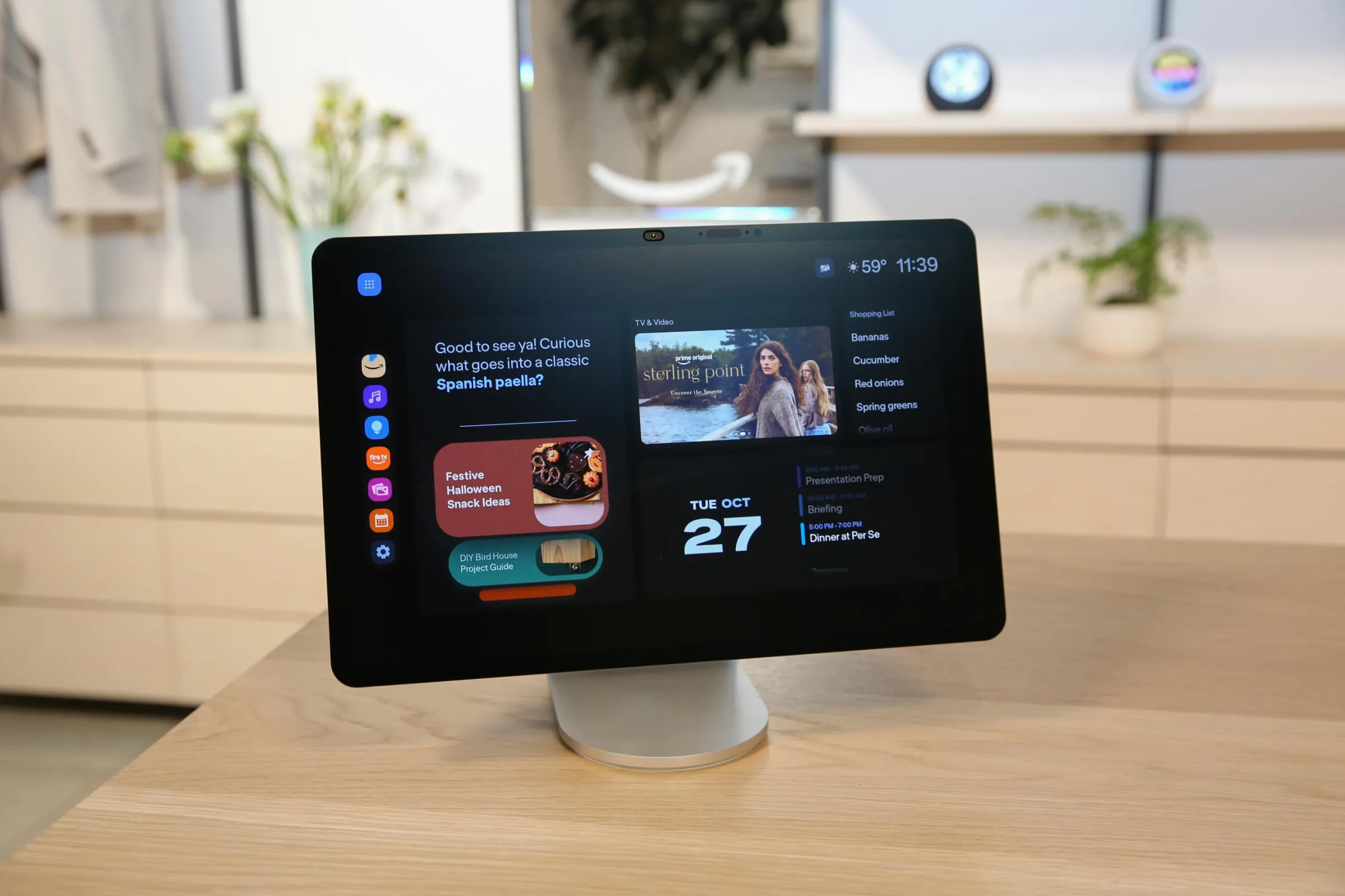 An Echo Show 16 is on a table in a kitchen. The display shows the date, app icons, a promo for a TV show, and more.