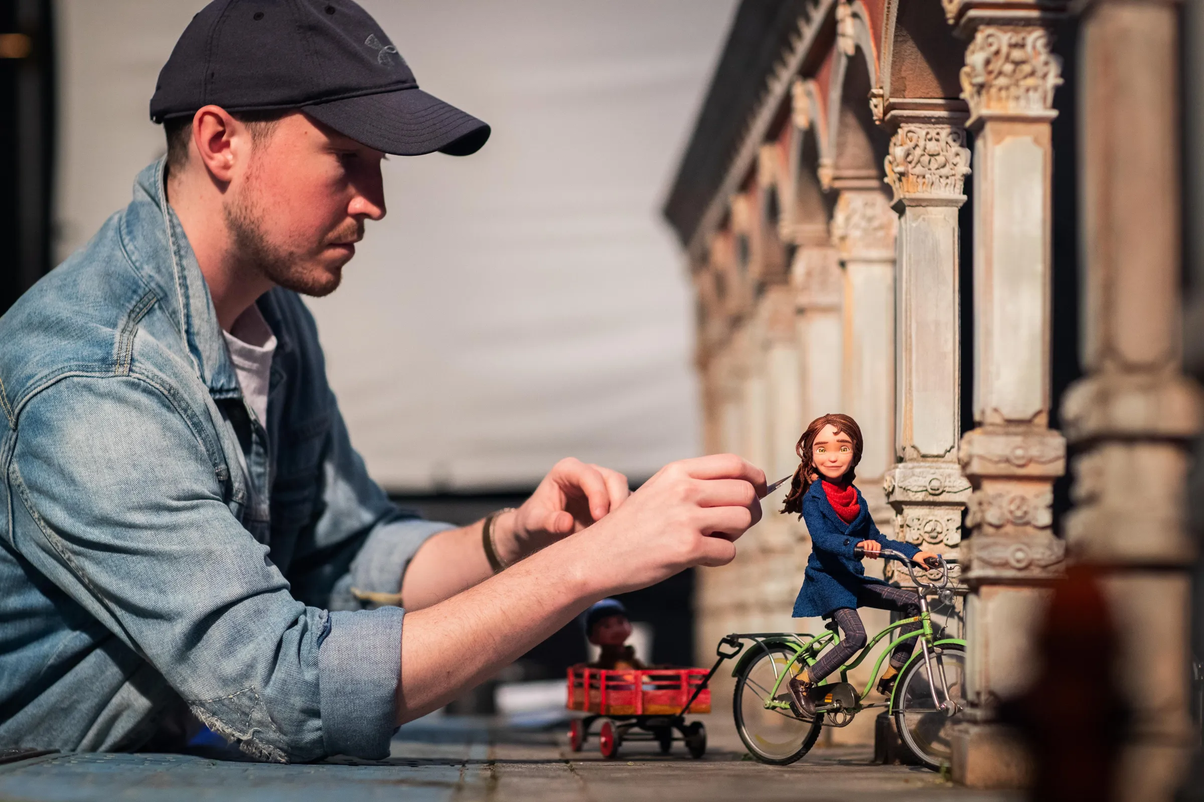 An animator uses a small tool on a puppet on a set for the stop-motion film Wildwood. The puppet is riding a bike near a building.
