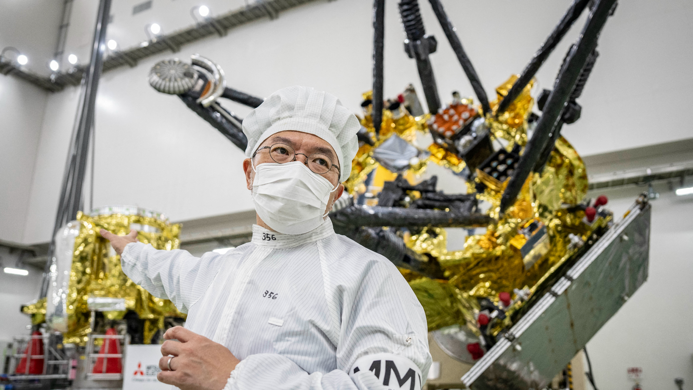 A photo of a man in white coveralls and a face mask stood in front two parts of the MMX probe in a clean room