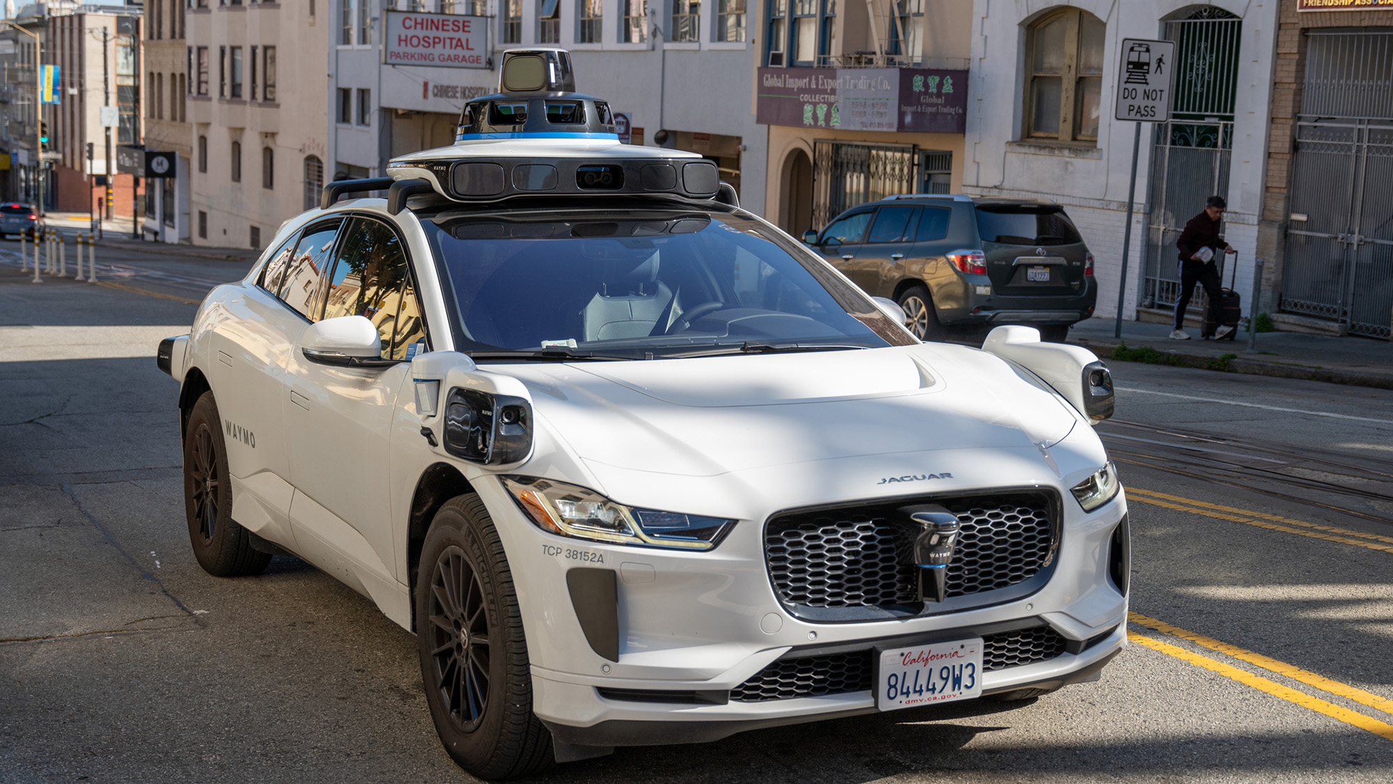 A white car with a camera on top is seen on a city street.
