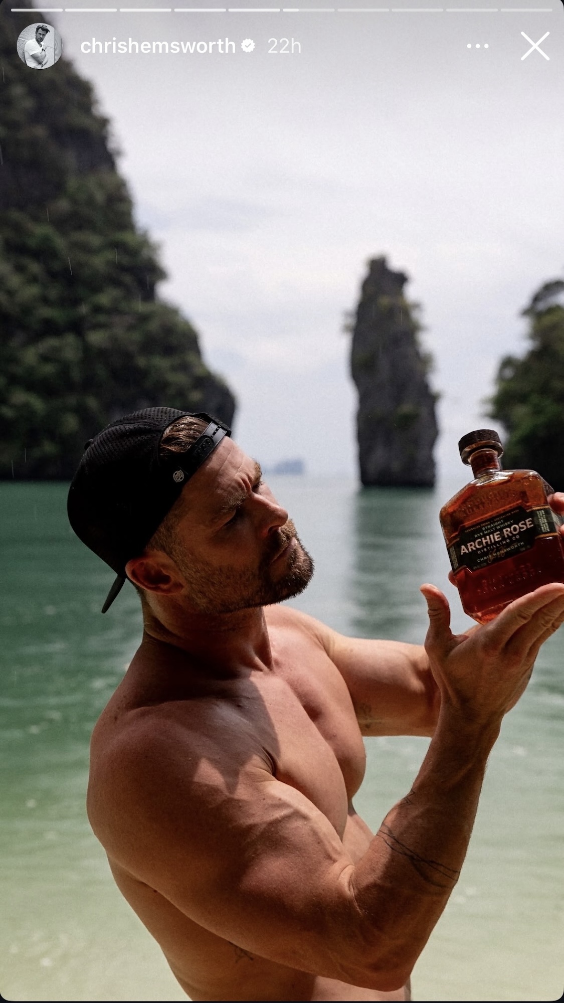 Shirtless Chris Hemworth wearing a hat and holding a bottle of Archie Rose from IG Story.