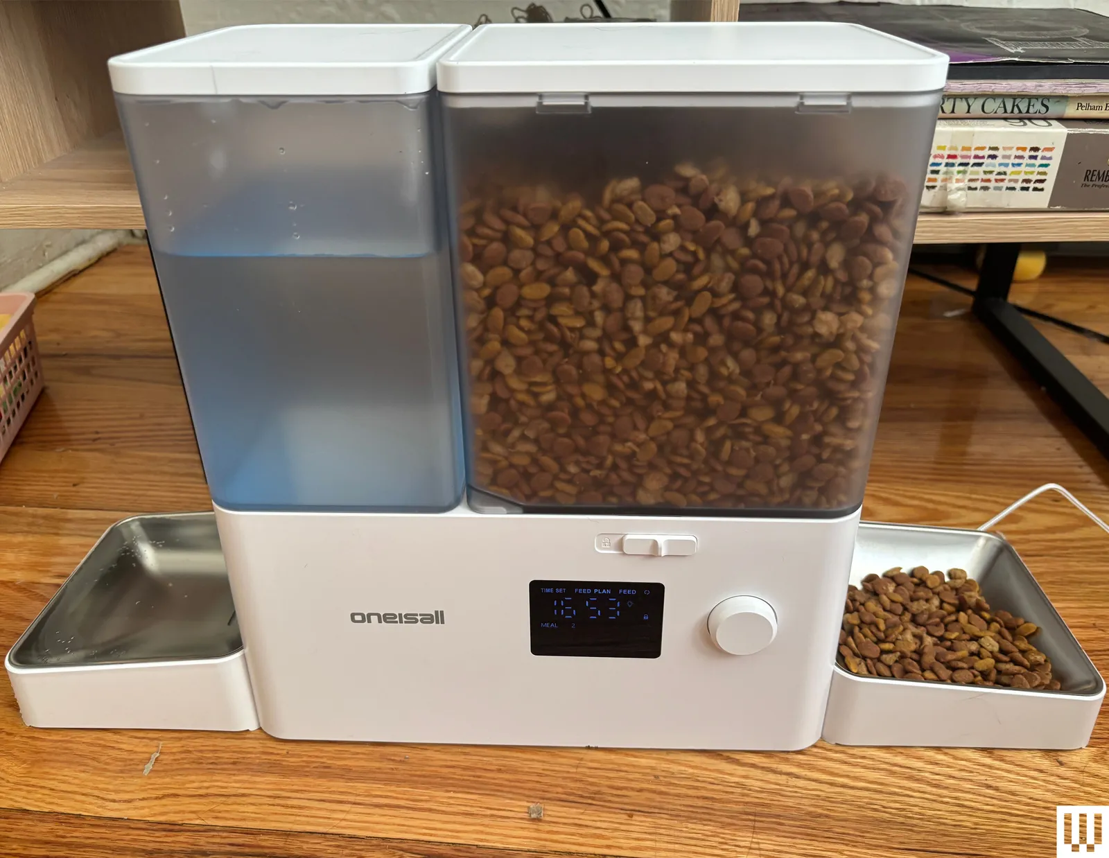 A white and clear automatic feeder and water dispenser for cats with brown kibble on a wood floor.