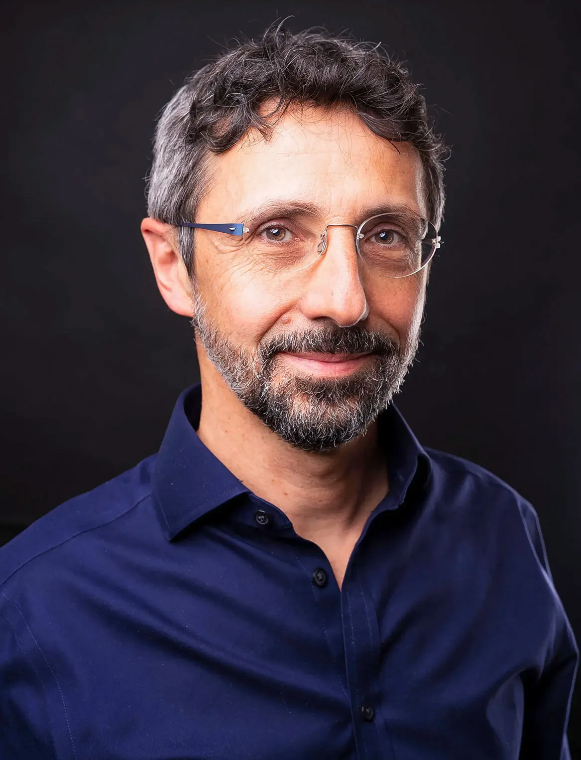 Headshot of a man with graying hair and a black beard with bits of gray. He wears rimless glasses and a dark blue button...
