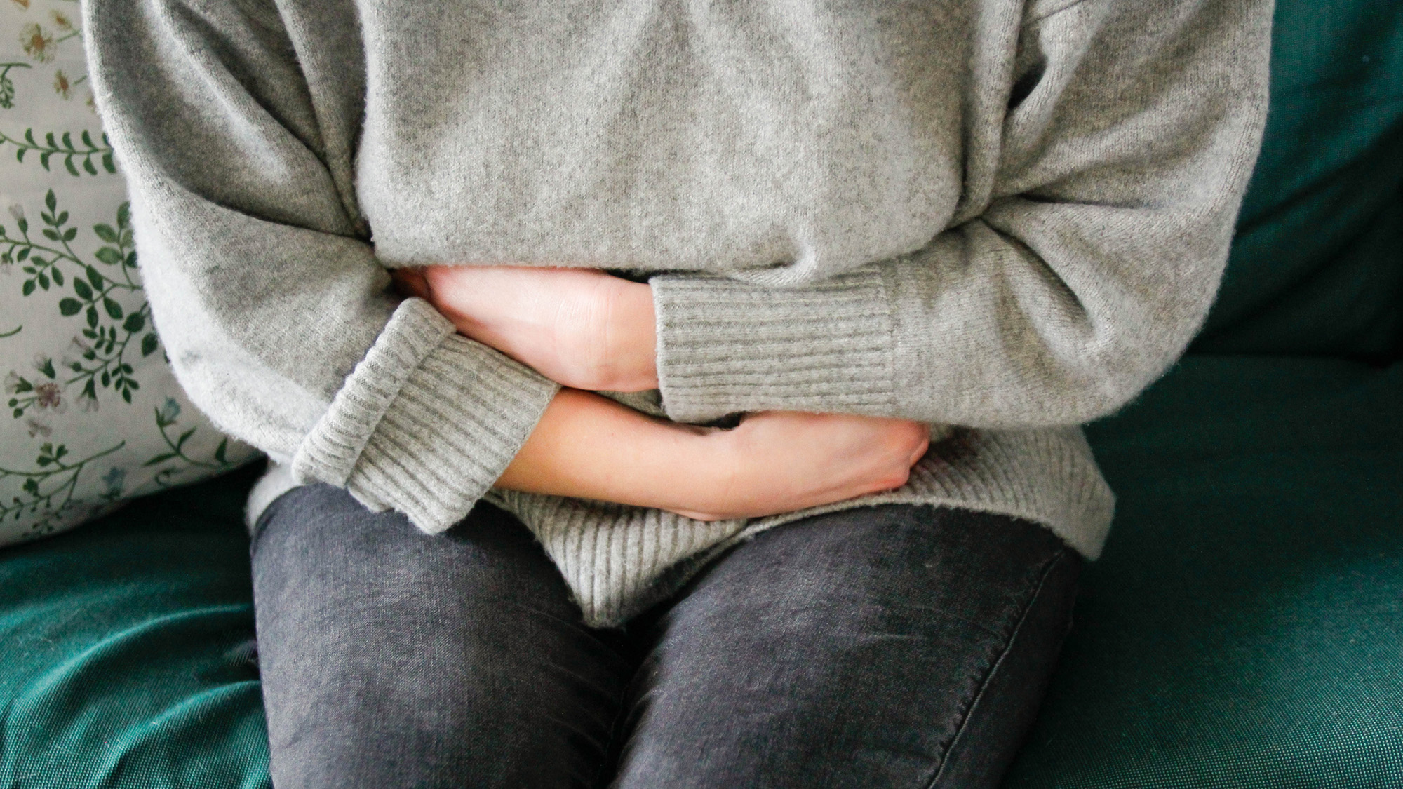 A woman wearing a gray sweater and blue jeans clutches her stomach.