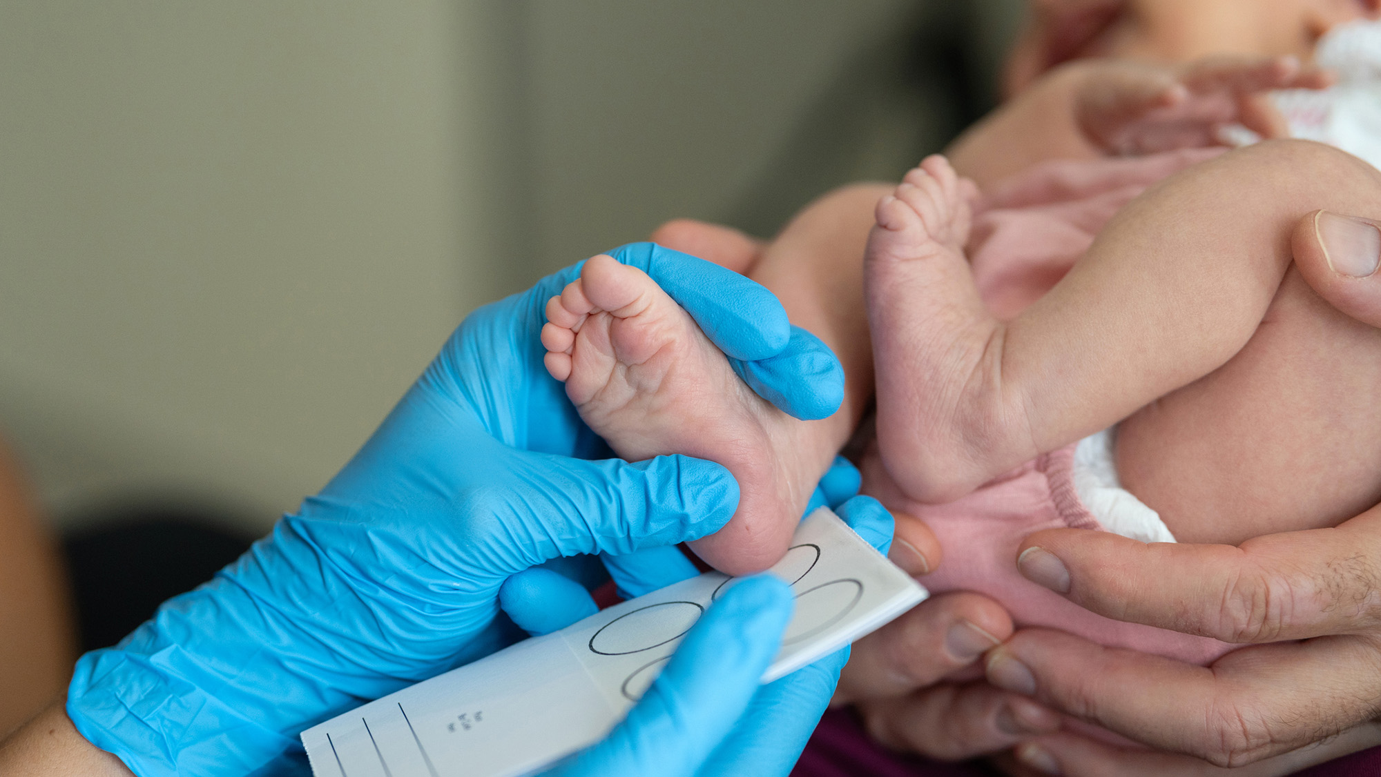 A blue gloved hand measures an infant's foot.