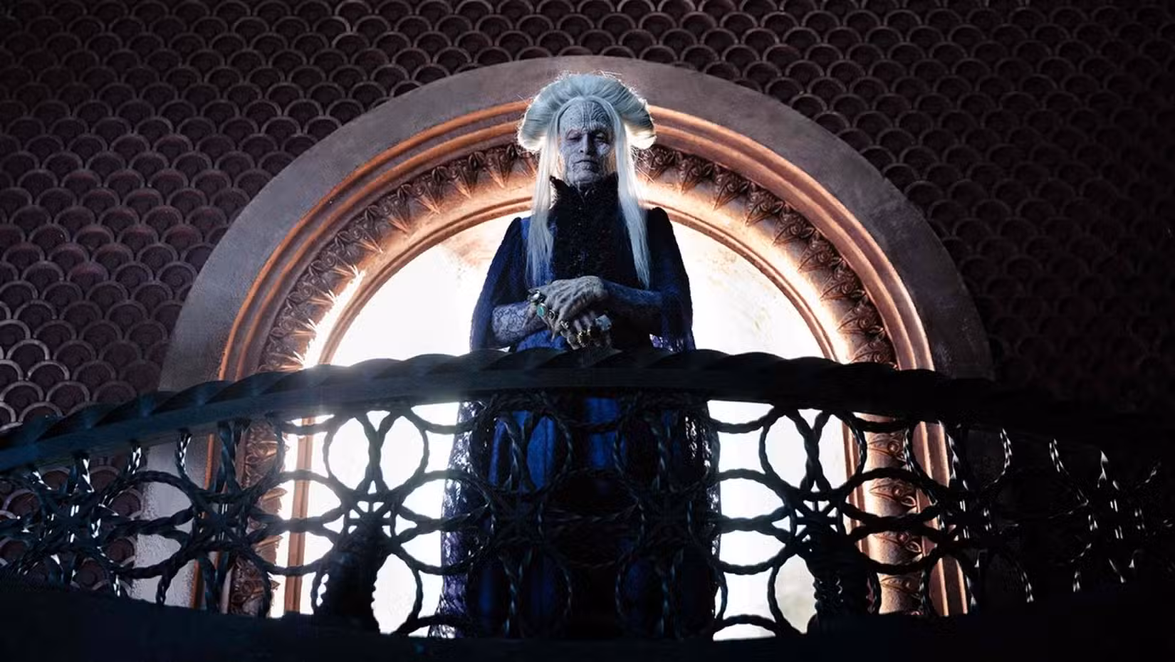In a scene from Luc Besson's Dracula, Caleb Landry Jones, playing the title monster, stands on an ornate balcony in front of a window, wearing his white hair 