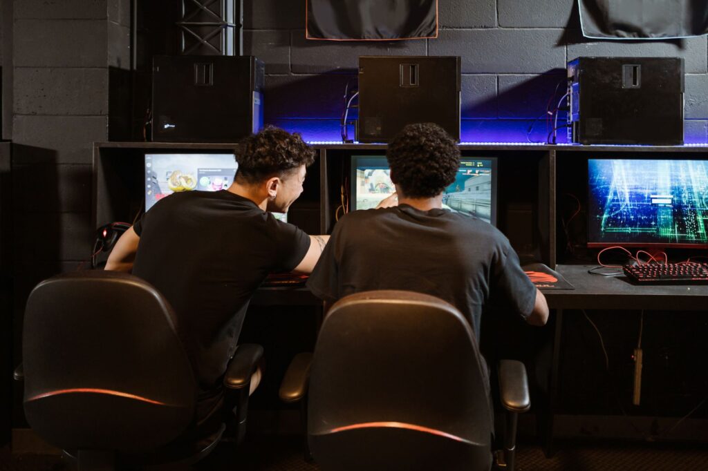 Two players seen from behind at gaming PCs with monitors in a dim esports room