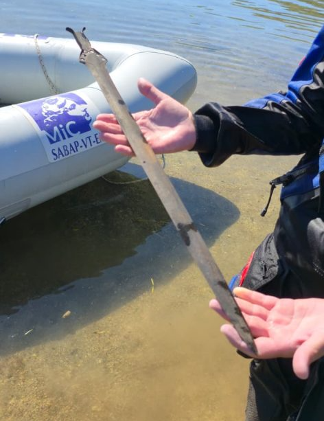 a person with light skin wears a dive suit and holds a Bronze Age sword, revealing its entire length; there is a boat in the background