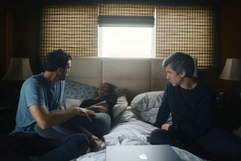 two men sitting on either edge of a bed while a blonde woman lies tearfully on her back.