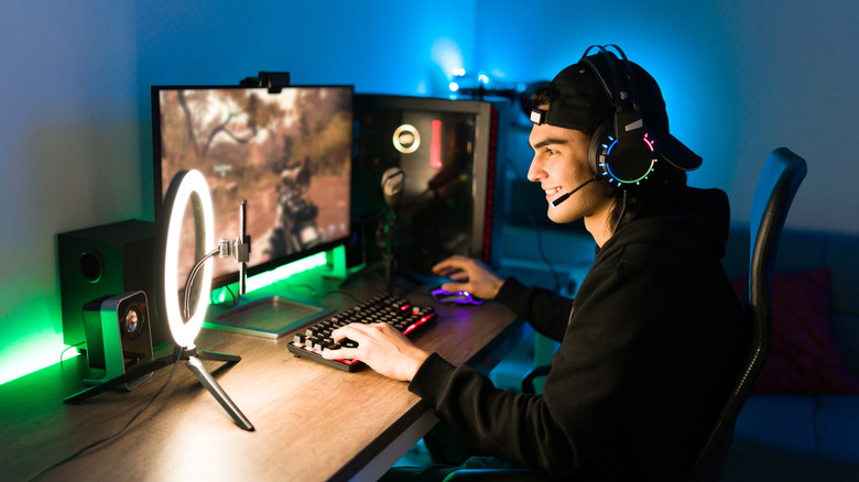 A teenage boy playing a first-person shooter on a gaming PC in a darkened room with accessories on the desk.