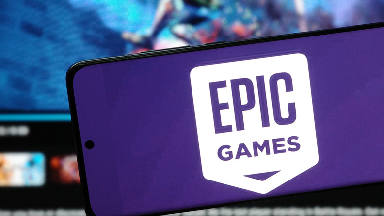 Photo of a smartphone displaying the Epic Games logo on a purple backdrop in front of a screen showing the company's website.