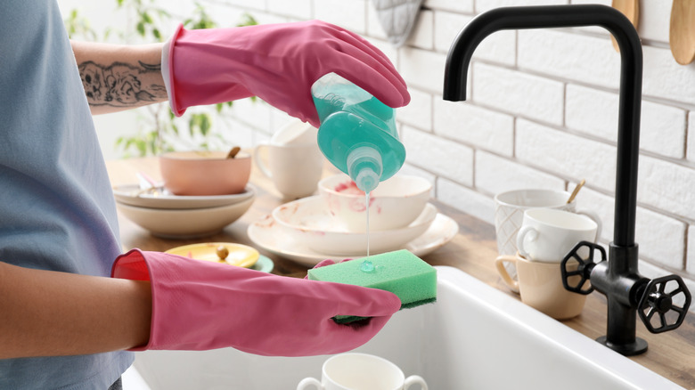 Close-up photo of a person wearing pink rubber gloves and squeezing dish soap onto a sponge. Dishes are stacked on the counter beside a farmhouse-style kitchen sink.