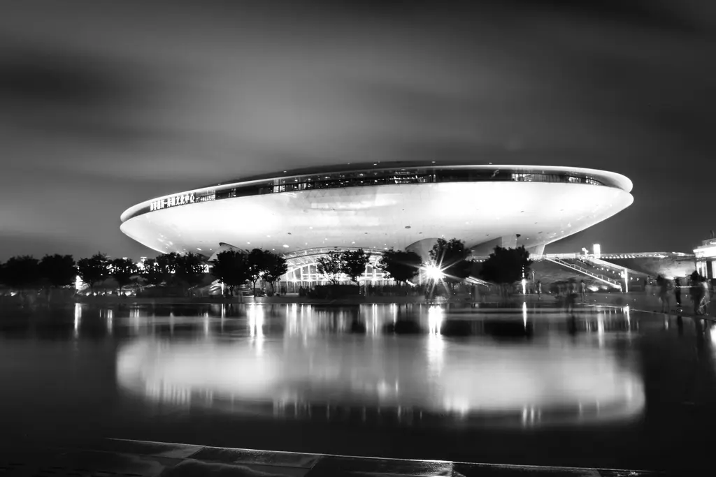 A black and white night view of the illuminated Mercedes-Benz Arena in Shanghai, reflected in a body of water.