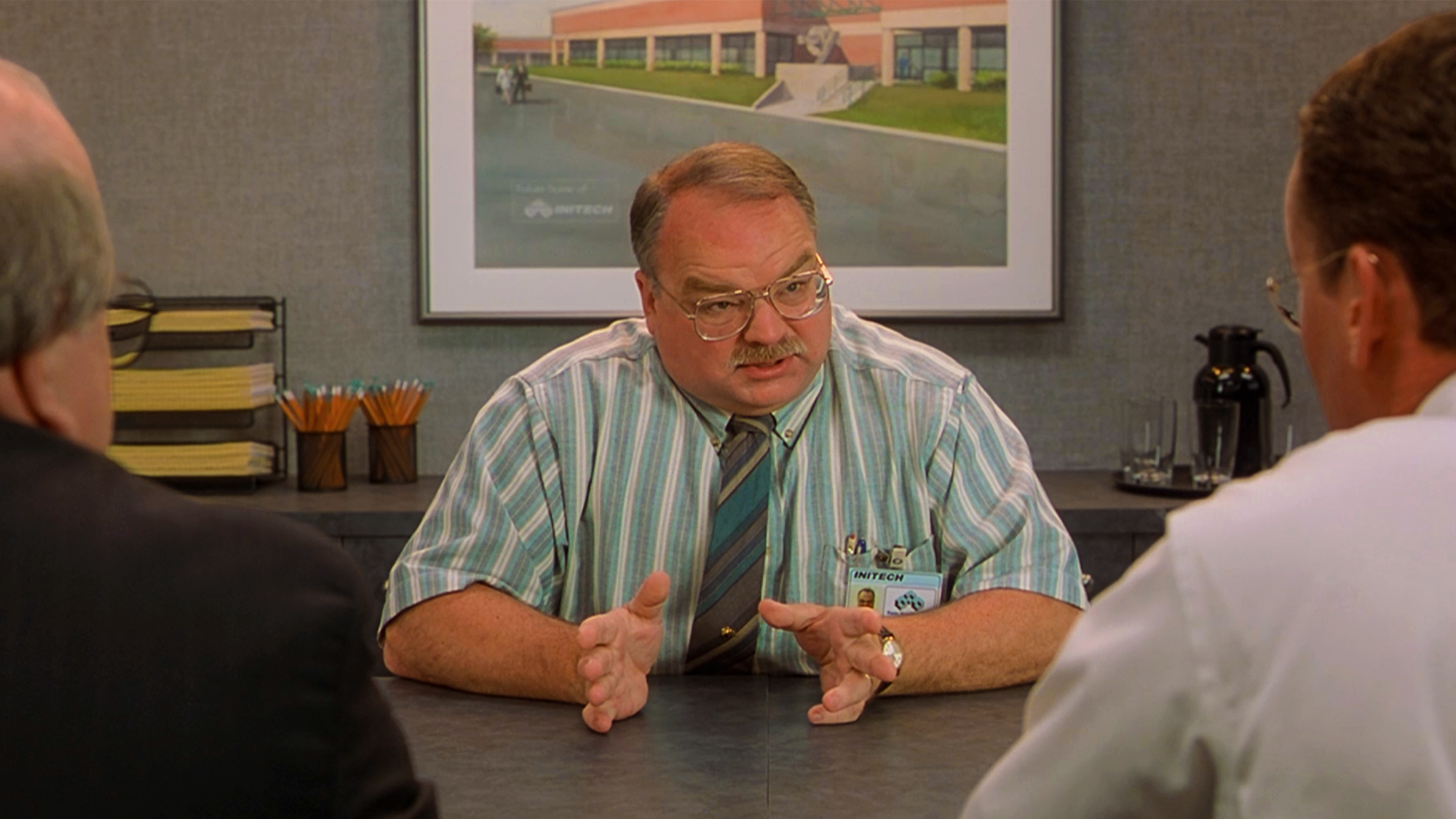 Riehle, dressed in a striped teal shirt and tie, wearing glasses, trying to explain what he 