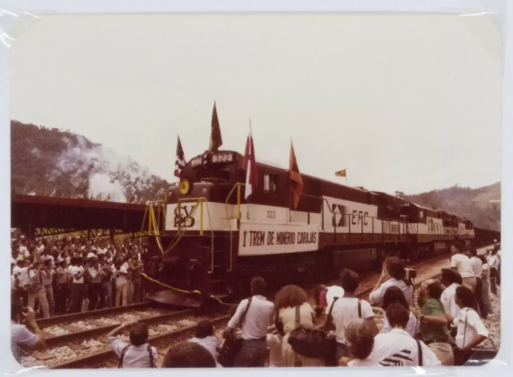 Uma multidão se reúne para observar um trem da linha Trem Carajás, enfeitado com bandeiras e uma faixa, parado nos trilhos.