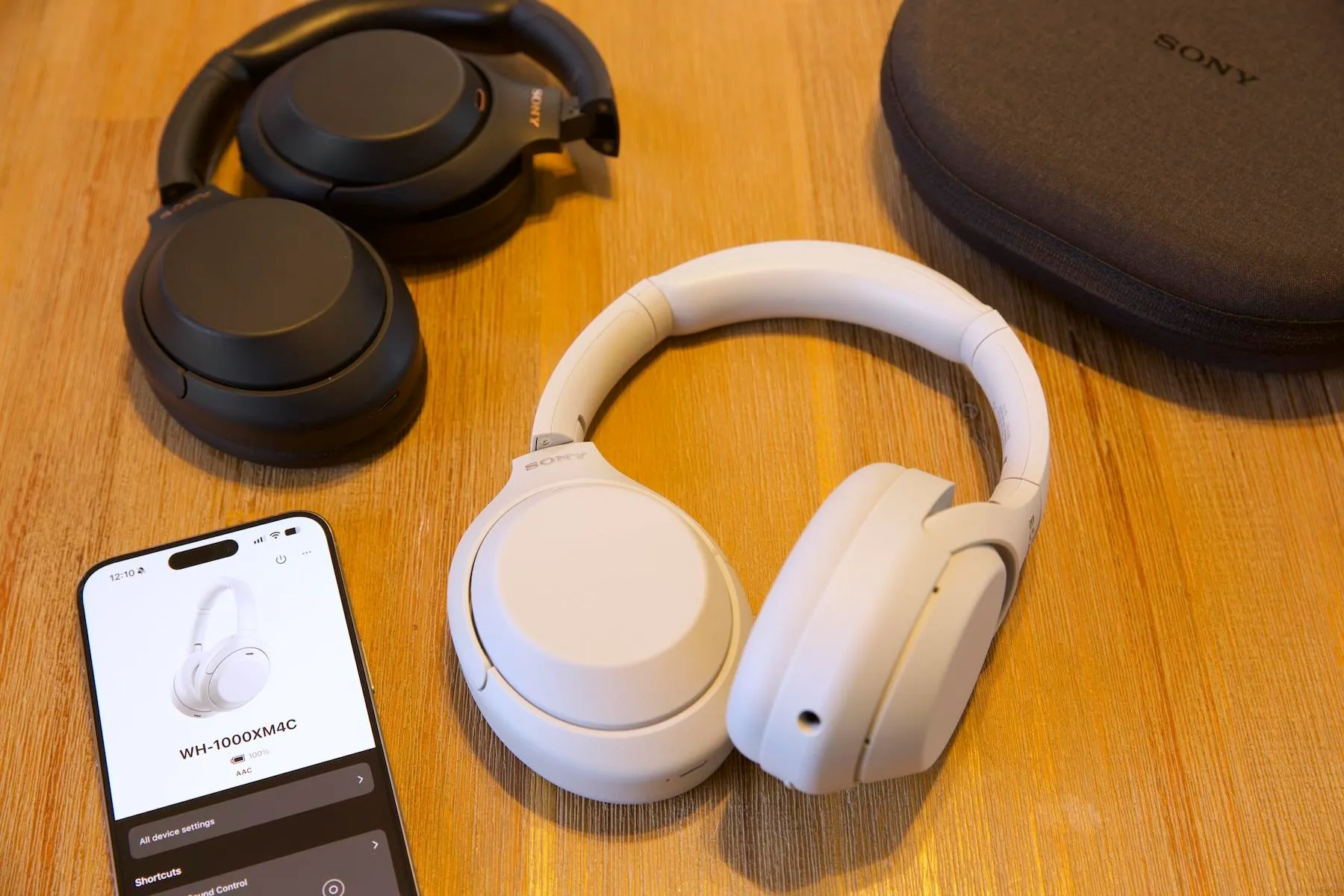 On a wooden table, a pair of white Sony WH-1000XM4C headphones are shown with the black WH-1000XM4 headphones along with an iPhone and a black carrying case.