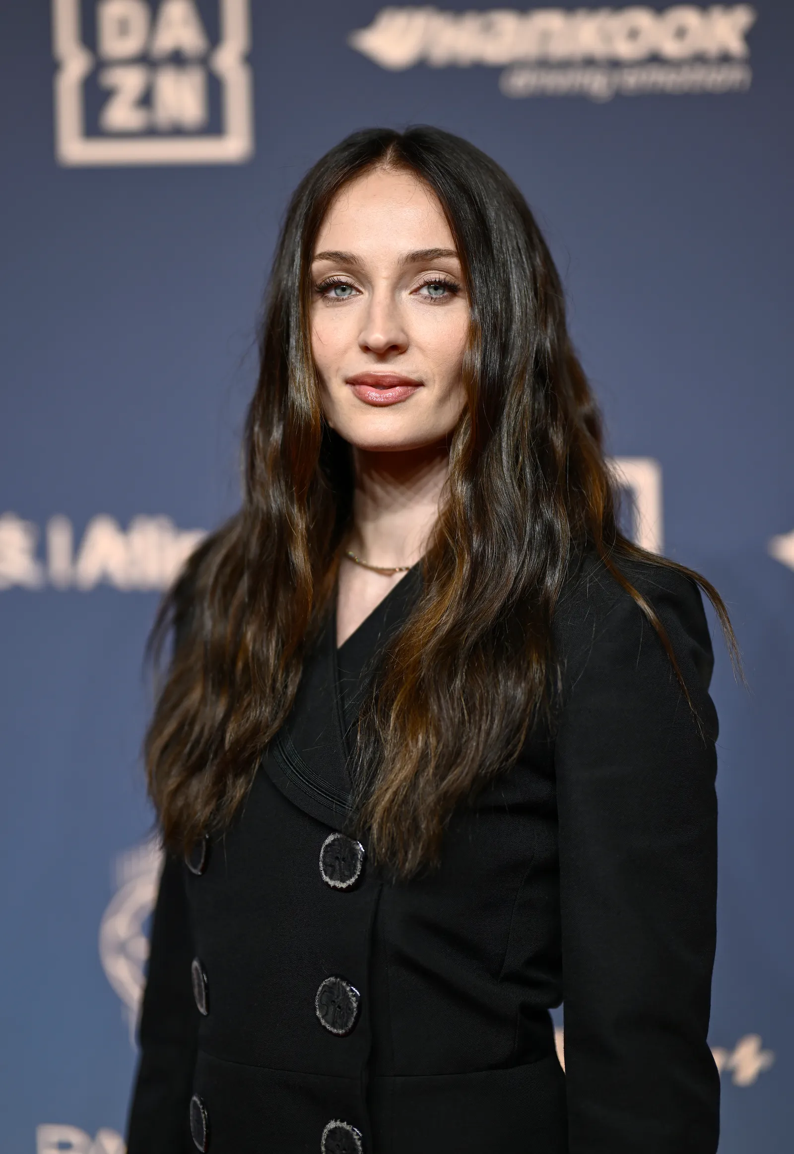 Sophie Turner wears a black jacket. She has long wavy dark brunette hair with highlights.