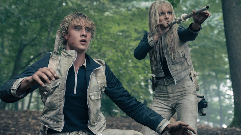 Joseph Zada as Haymitch kneeling by McKenna Grace as Maysilee holding her blowgun in The Hunger Games: Sunrise on the Reaping