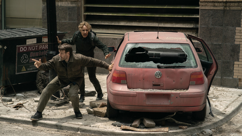 Joel Edgerton as Jason Dessen and Amanda Brugel as Blair Caplan crouching behind a destroyed car in a war-torn street in Dark Matter
