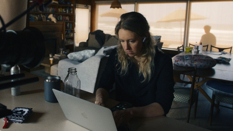 Elizabeth Holmes working on her laptop with Nathan Fielder in the backround outside the window in You Can See Everything